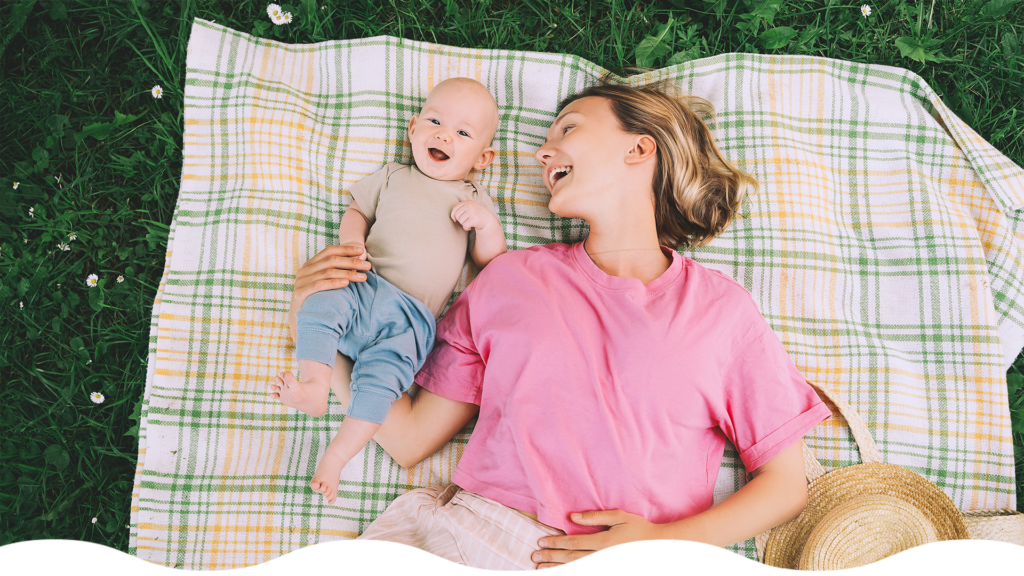 Mamá sonriente y bebé descansando juntos sobre una manta en el pasto, disfrutando de actividades sensoriales para bebés en la naturaleza